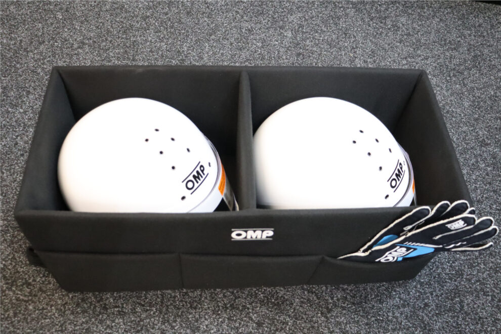 Two white helmets sitting in the two compartments of a black OMP helmet box, with gloves in a side pocket