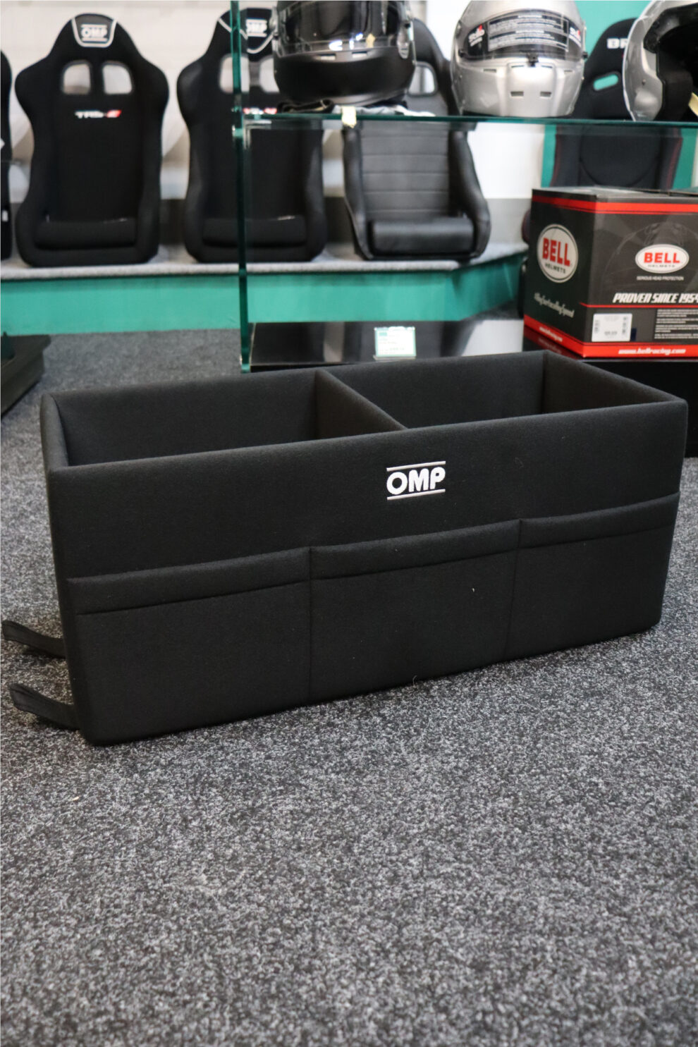 An open black OMP double helmet box with two compartments and pockets on the front, on the carpet in the GSM Performance showroom with seats and helmets behind