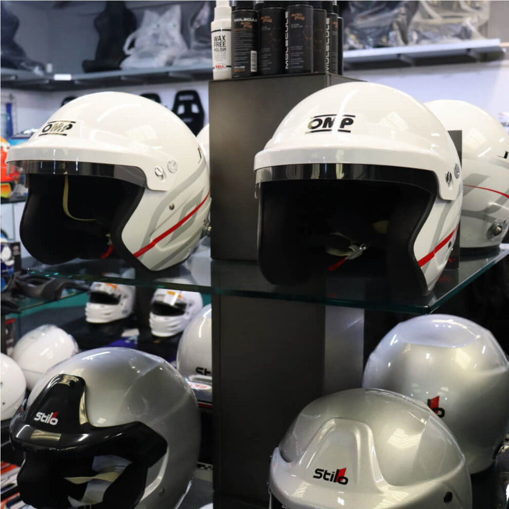 Two white OMP J-R open face helmets with red and grey graphics on a glass shelf in the GSM Performance showroom, with Stilo helmets below