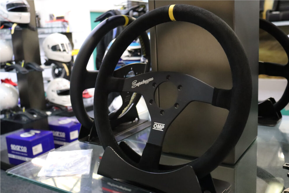 A black suede OMP Velocita Superleggero steering wheel on a display stand in the GSM Performance showroom, with helmets on shelves behind