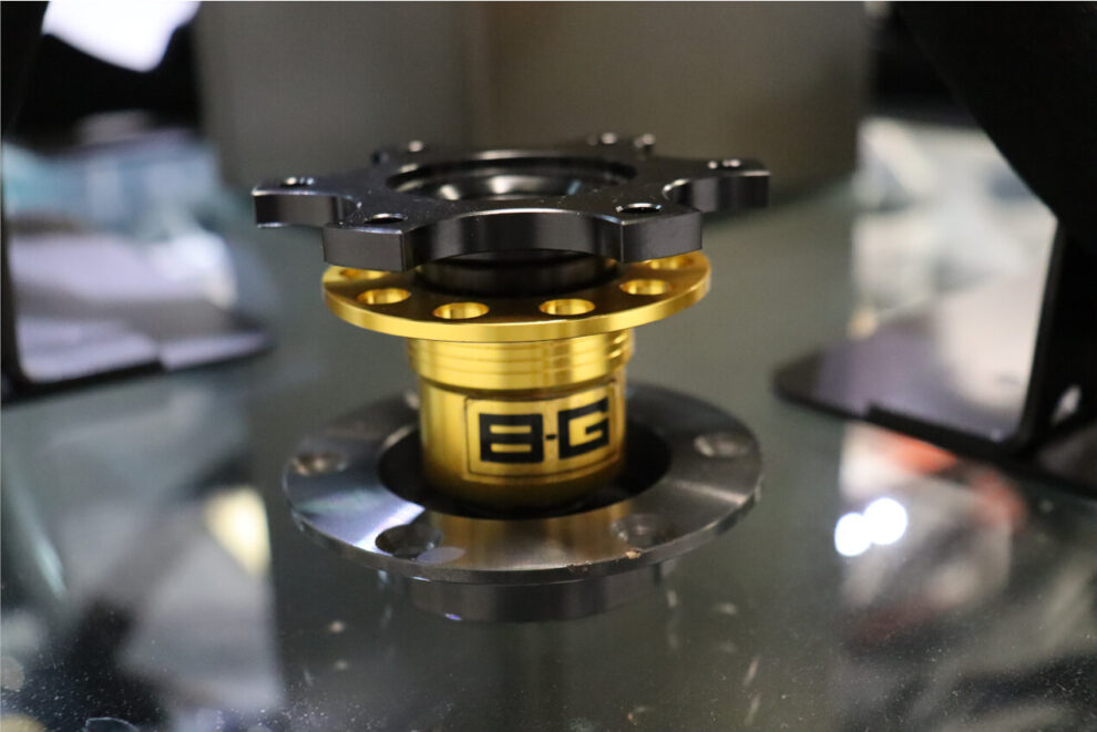 A gold and black B-G Racing quick release steering wheel boss on a glass shelf