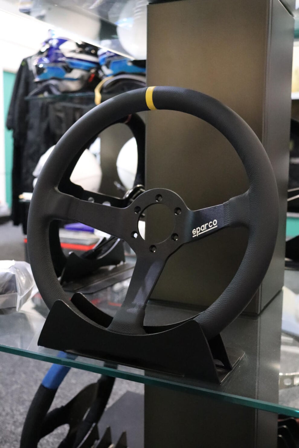 Sparco 345 steering wheel on a display stand on a glass shelf with race suits behind