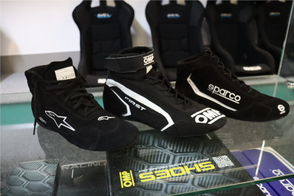 Alpinestars SP V2, OMP First and Sparco Slalom race boots side by side on a glass shelf in the GSM Performance showroom, with bucket seats behind