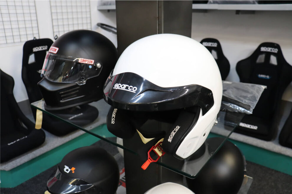 A white Sparco open face helmet on a glass display stand, with a black Simpson helmet and Sparco bucket seats behind