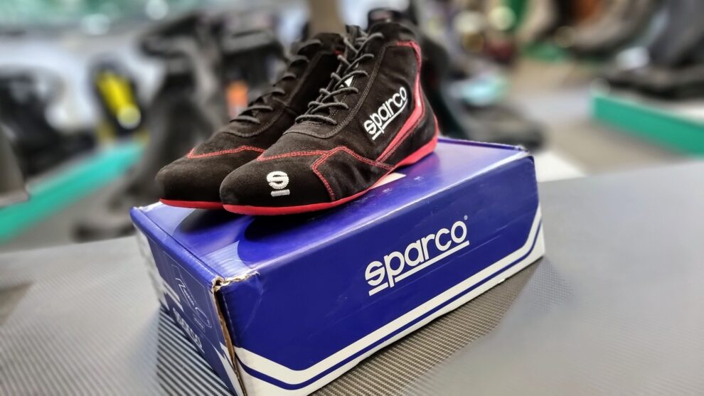 A pair of black suede Sparco Slalom racing shoes with red stitching and red soles standing on their blue Sparco box in the GSM Performance showroom
