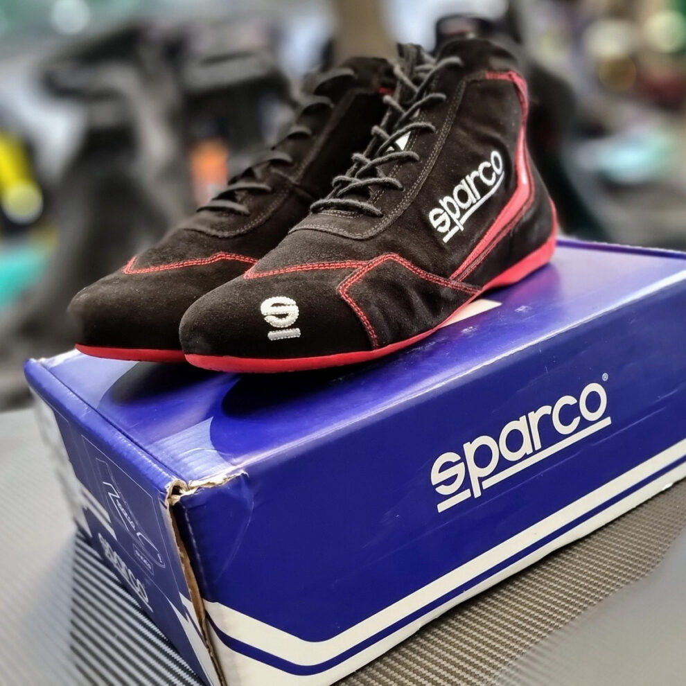 Black suede Sparco Slalom racing shoes with red stitching on a blue Sparco box, with racing seats blurred behind