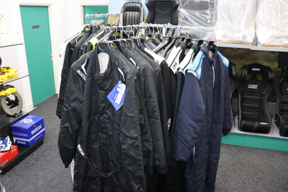 A rail of black Sparco and navy OMP race suits in the GSM Performance showroom, with bucket seats on the shelves behind