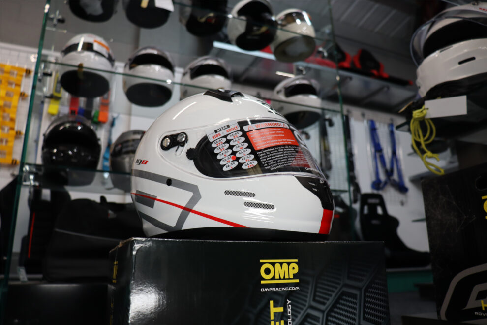 A white OMP KJ-8 Evo karting helmet with grey and red graphics on its box in the GSM Performance showroom, with shelves of helmets behind