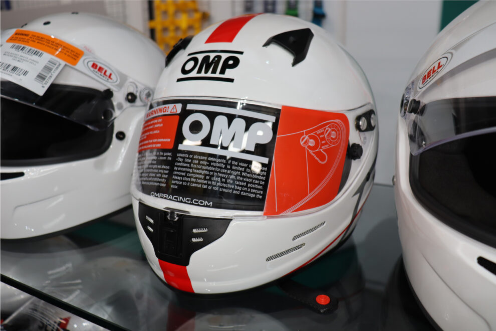 A white OMP karting helmet with an orange visor sticker and red stripe on a glass shelf between two white Bell helmets