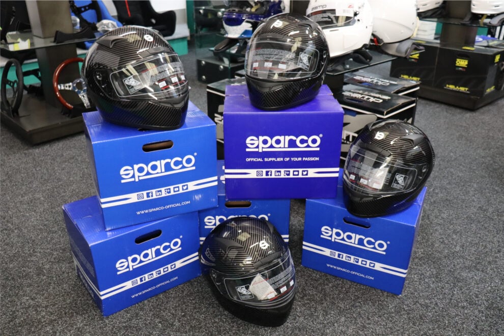 Sparco Club X1 Carbon helmets stacked on blue and purple Sparco boxes in front of the helmet wall
