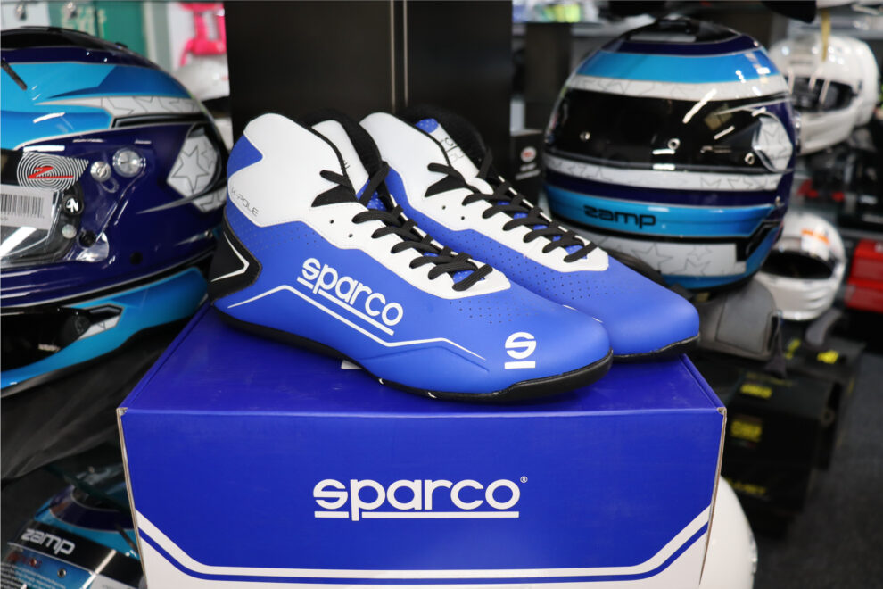 A blue and white Sparco K-Pole kart boot on its box, with race helmets on the shelf behind
