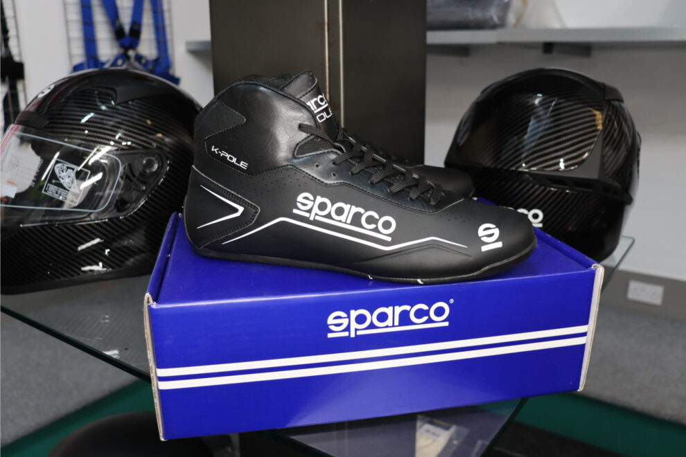 A black Sparco K-Pole kart boot on its box between two carbon helmets in the showroom