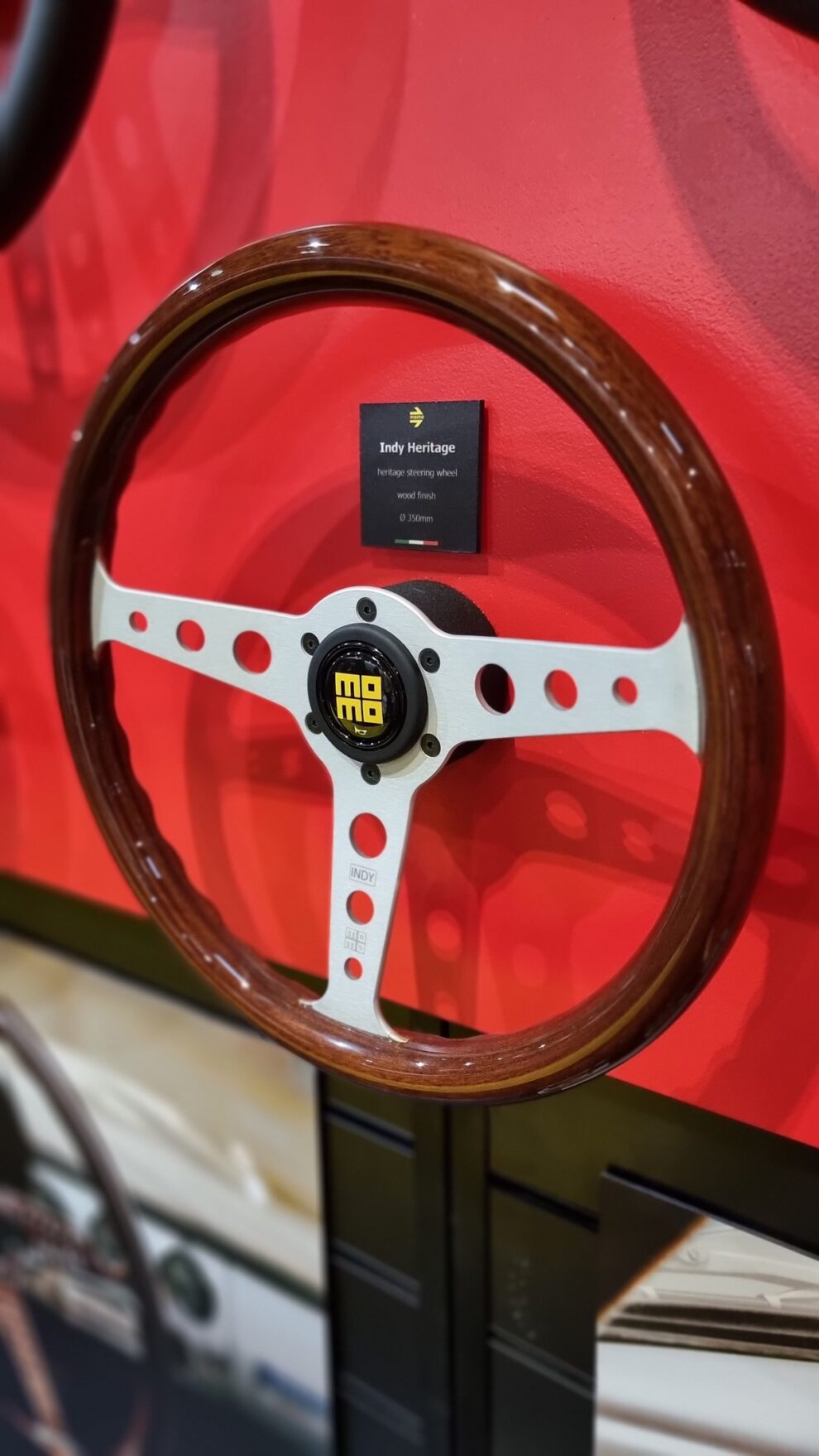 MOMO Indy Heritage steering wheel with a wooden rim and drilled silver spokes on the MOMO stand