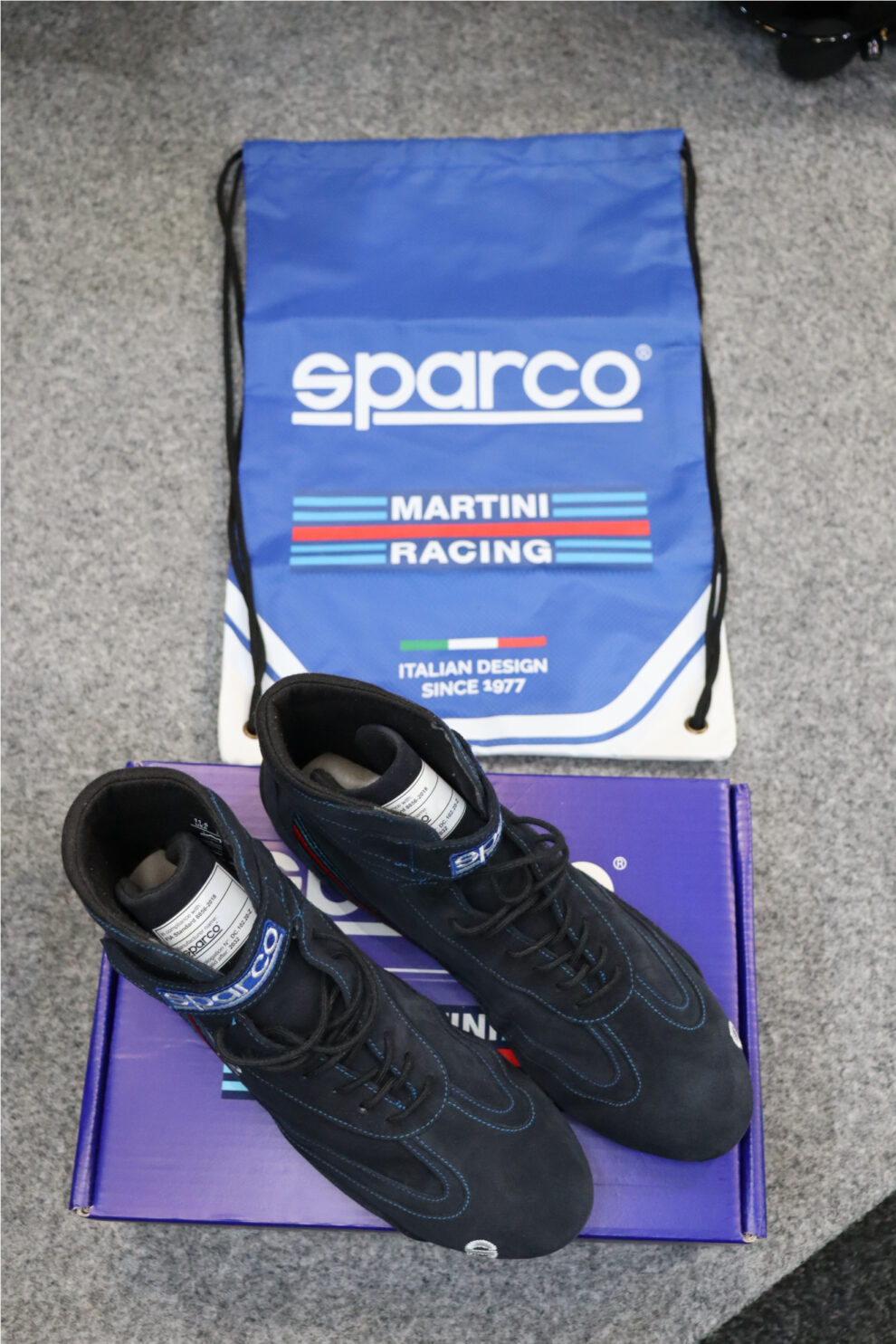 Sparco Martini Racing boots on their box beside a blue Sparco Martini Racing drawstring bag