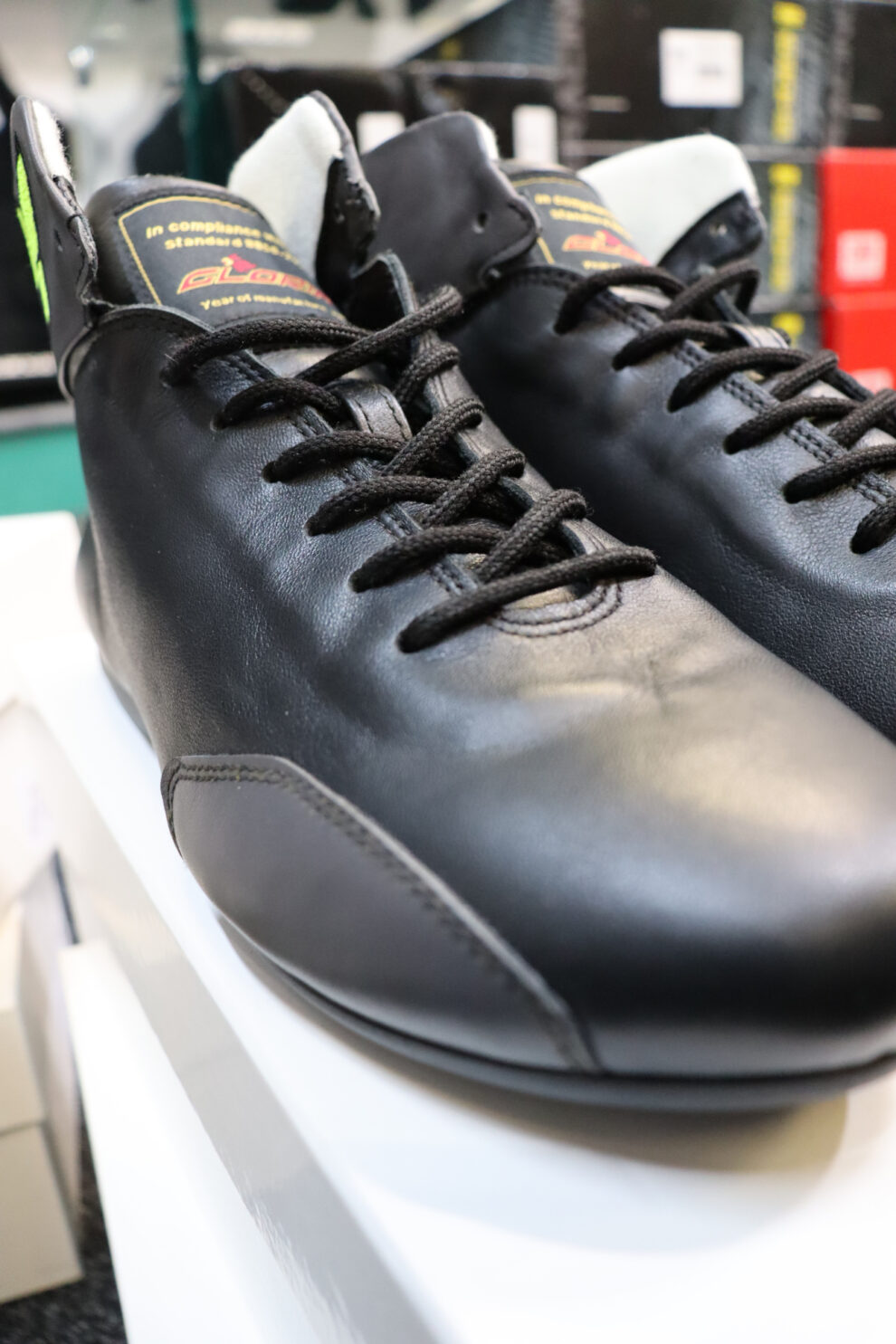 A close view of a pair of black leather P1 Monza racing boots with black laces on a white box