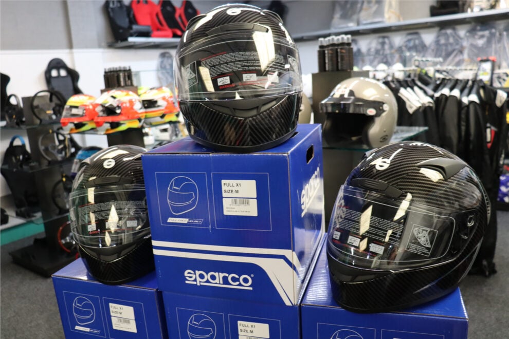 Three Sparco Club X1 Carbon helmets on Sparco boxes in the showroom, with coloured helmets and seats behind