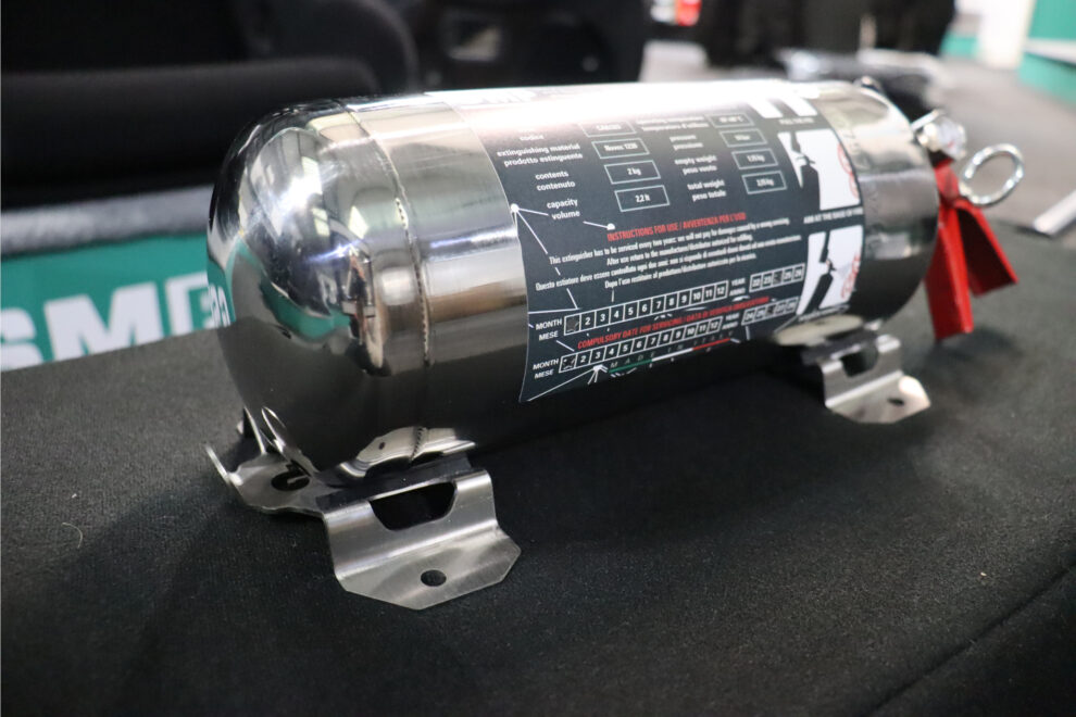A polished stainless steel OMP Novec 2 kg hand held fire extinguisher with its mounting brackets, on a black display plinth in the GSM Performance showroom