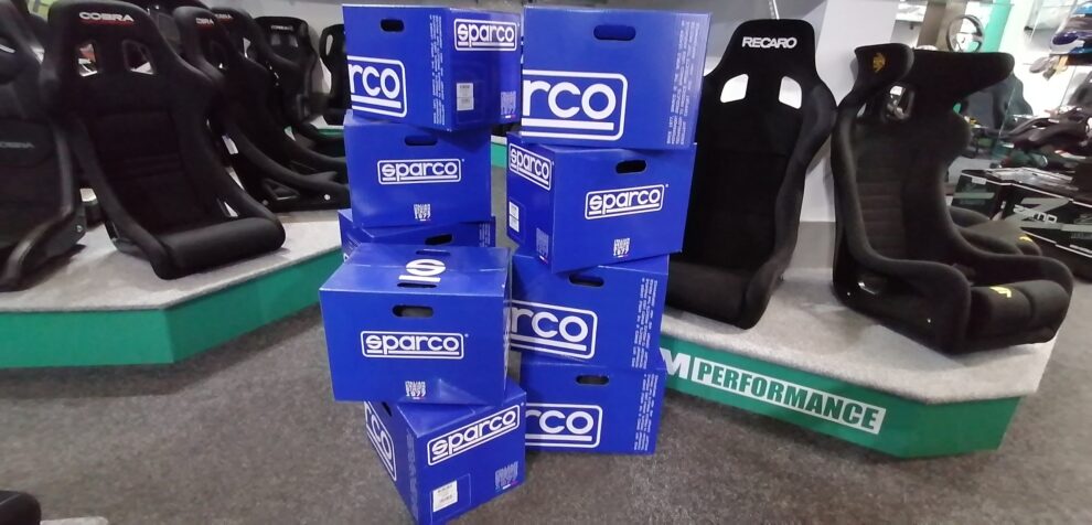 A stack of blue Sparco delivery boxes on the floor of the GSM Performance showroom, between displays of Cobra and Recaro bucket seats