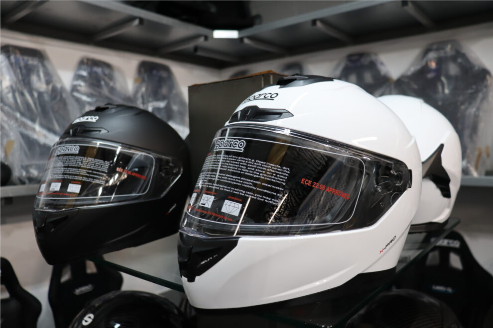 A matt black and a white Sparco X-Pro full face helmet on a glass shelf in the GSM Performance showroom, with an ECE 22.06 approved sticker on the white visor