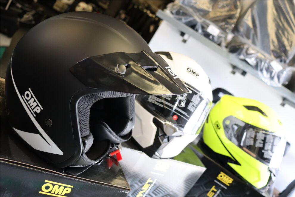 Close view of a matt black OMP Star open face helmet with a black peak, with white and fluro yellow Circuit Evo 2 helmets behind on OMP boxes