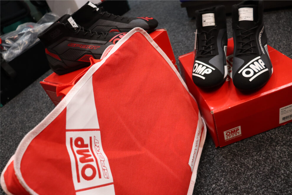 Black and white OMP Sport race boots on a red OMP box next to a red OMP Sport bag, with a black and red pair behind