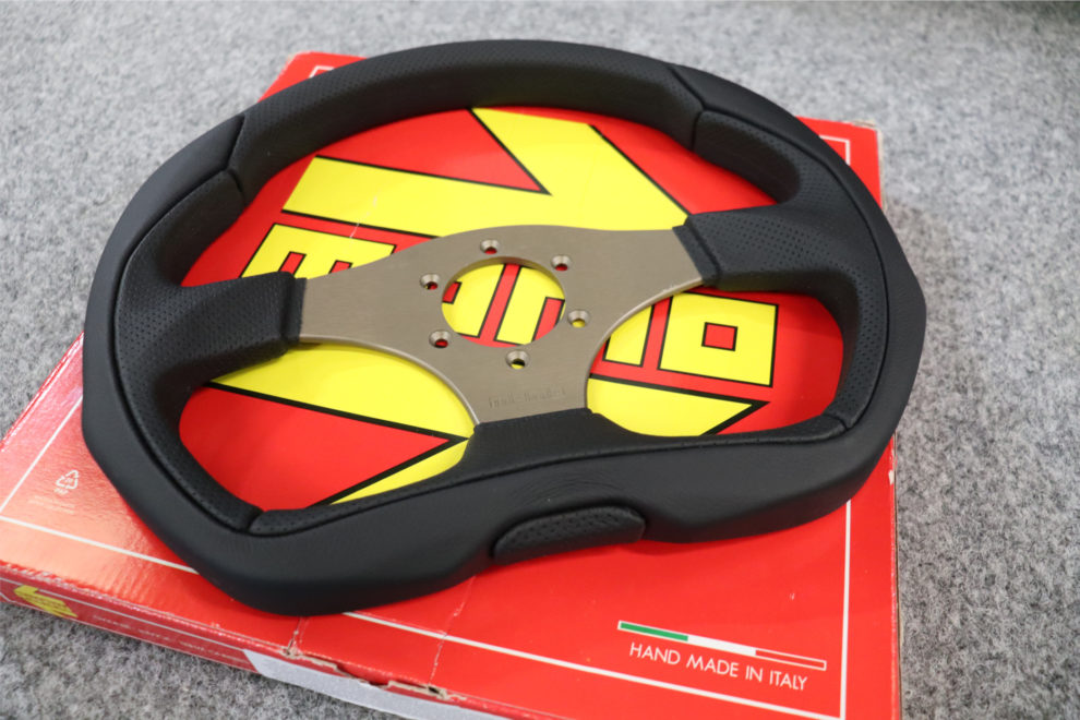 A MOMO Eagle flat bottom steering wheel in black leather with an anthracite spoke plate, resting on its red and yellow MOMO box