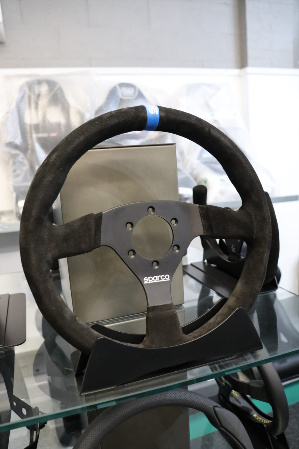 The Sparco 383 steering wheel with a blue logo stripe on a stand on a glass shelf, with seats in covers behind