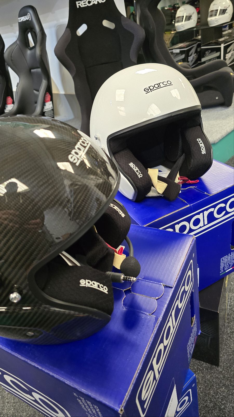 The Sparco RJ-i Carbon helmet with its microphone boom on a blue Sparco box, beside a white Sparco open face helmet, with Recaro and Corbeau seats behind