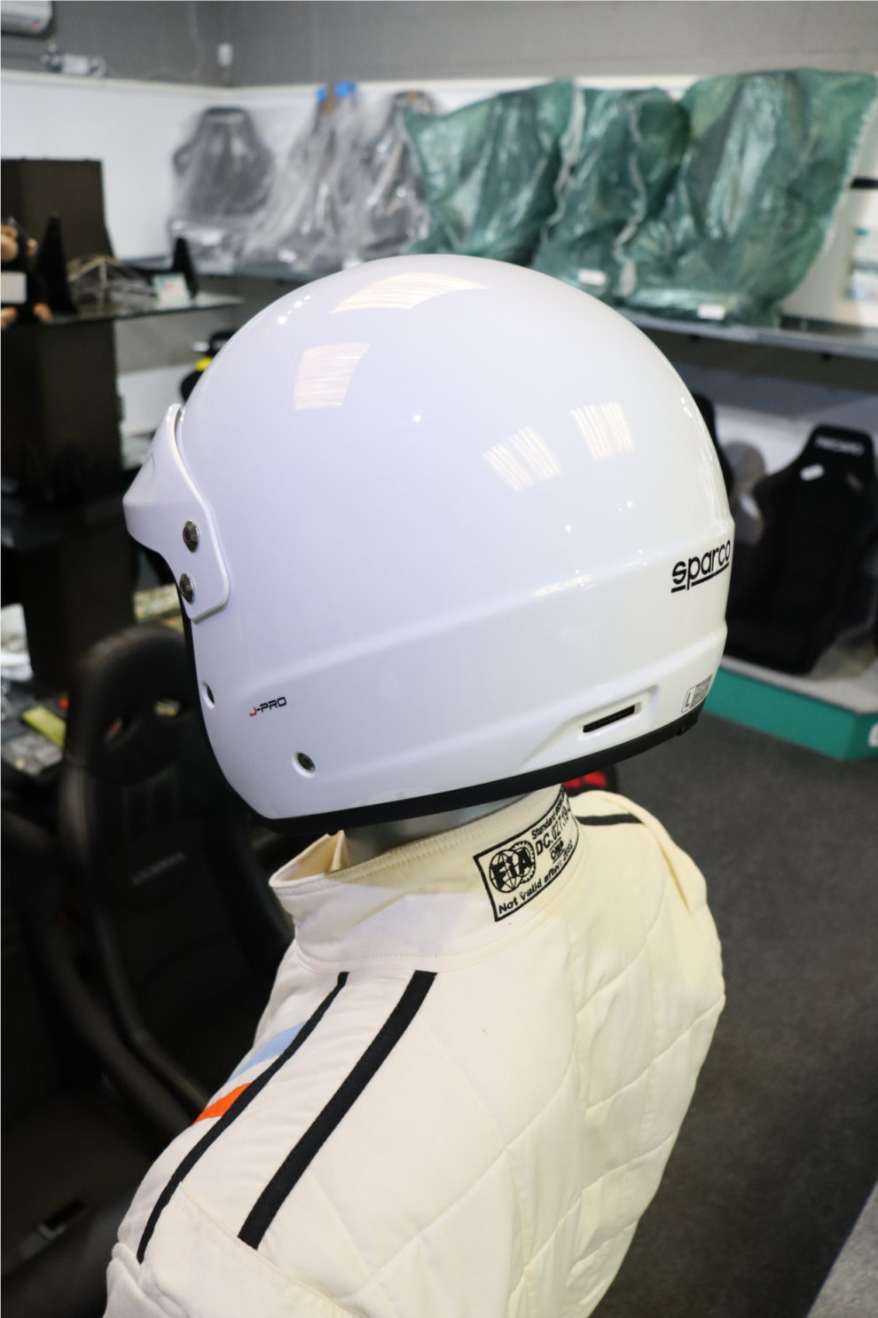 A white Sparco J-Pro helmet seen from the side on a mannequin wearing a race suit, in the GSM Performance showroom