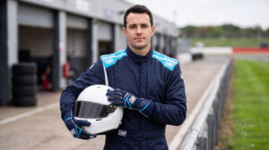 Driver in a blue OMP First race suit holding a correctly forward-facing white helmet with a dark visor in a UK circuit pit lane