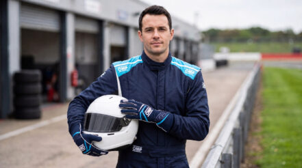 Driver in a blue OMP First race suit holding a correctly forward-facing white helmet with a dark visor in a UK circuit pit lane