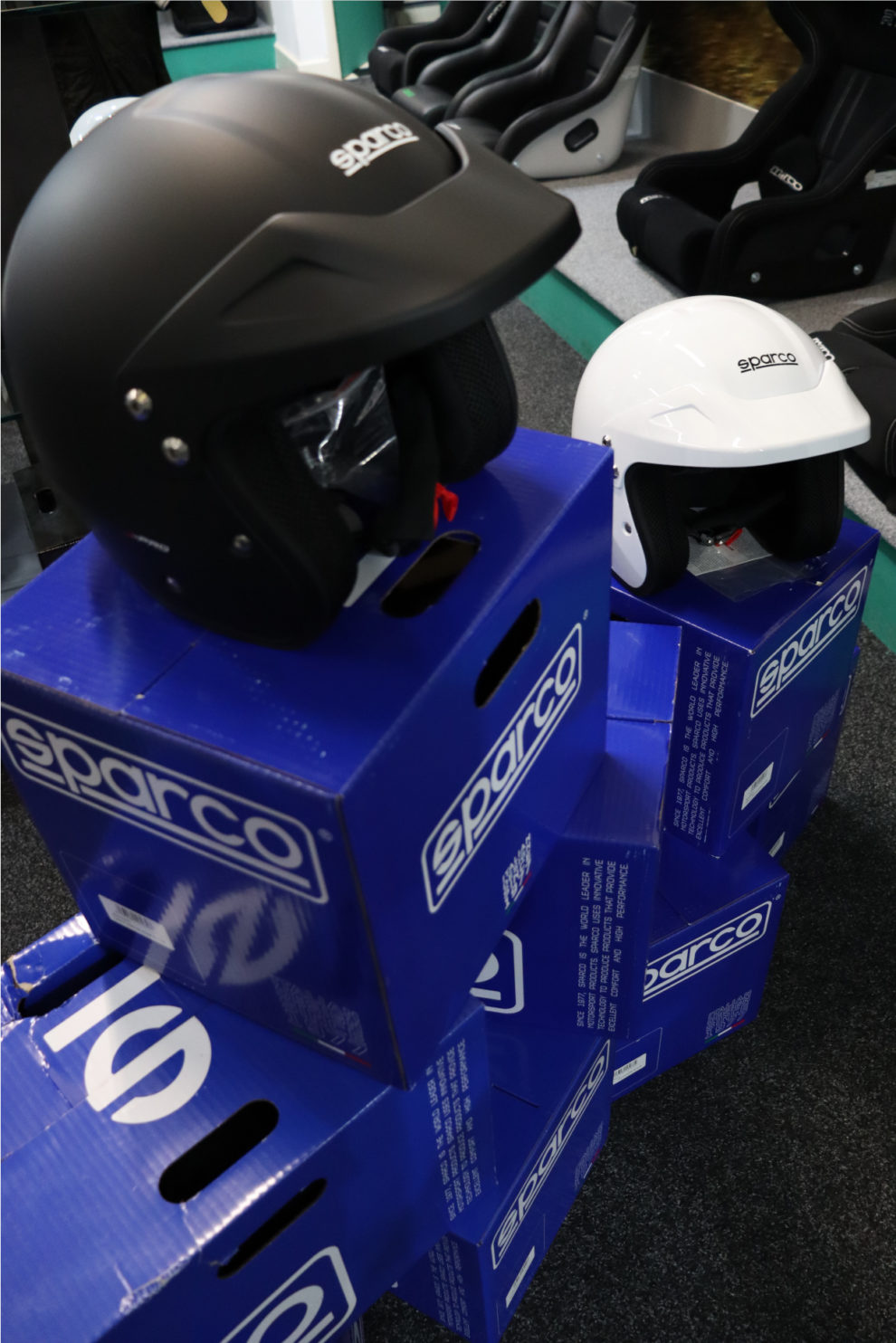 A matt black Sparco J-Pro with its peak and a white J-Pro behind it, both on blue Sparco boxes in the GSM Performance showroom with seats in the background