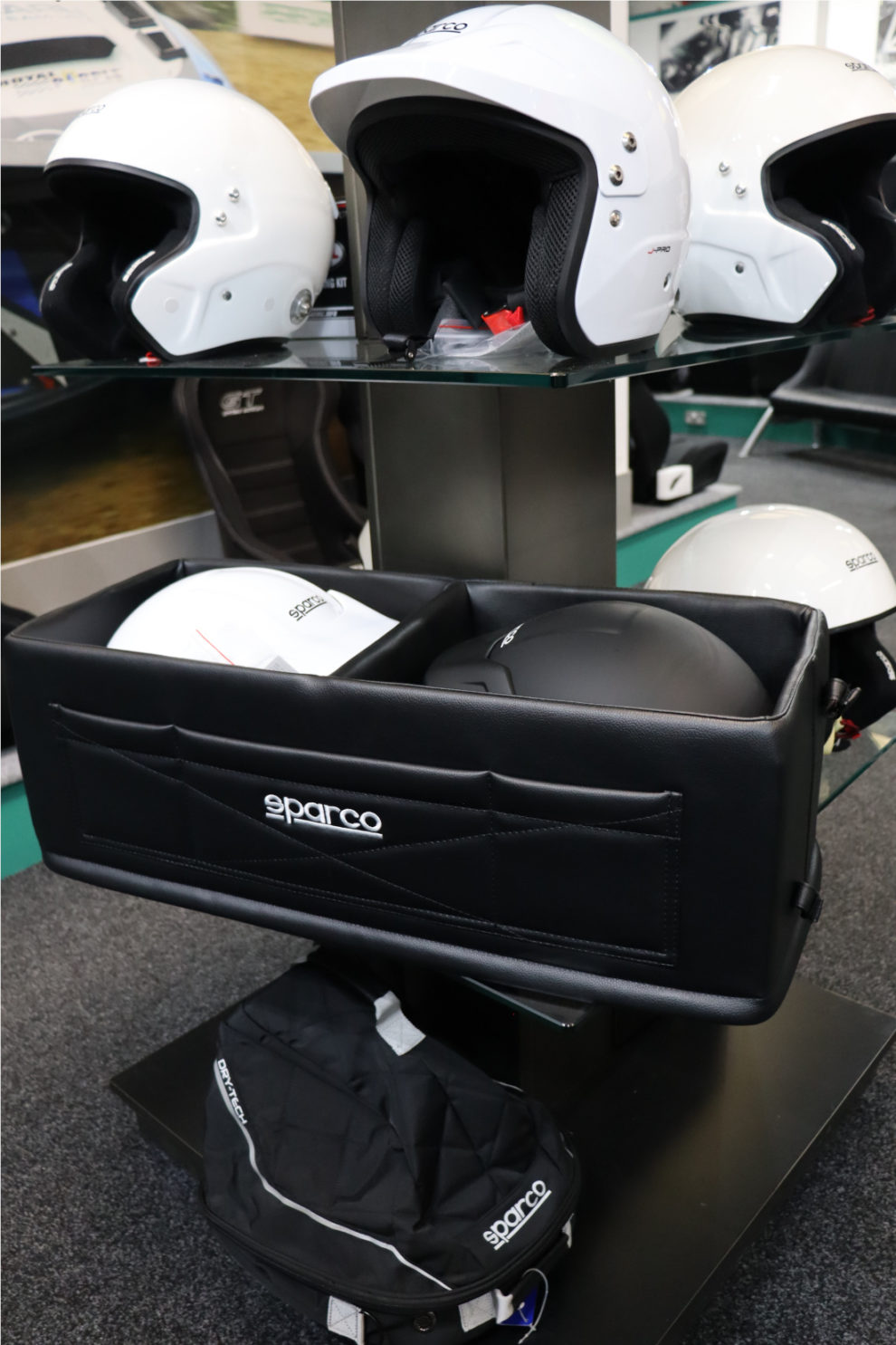 The black Sparco Double Helmet Box on a showroom display stand, with white open face helmets on the glass shelf above and a Sparco bag below