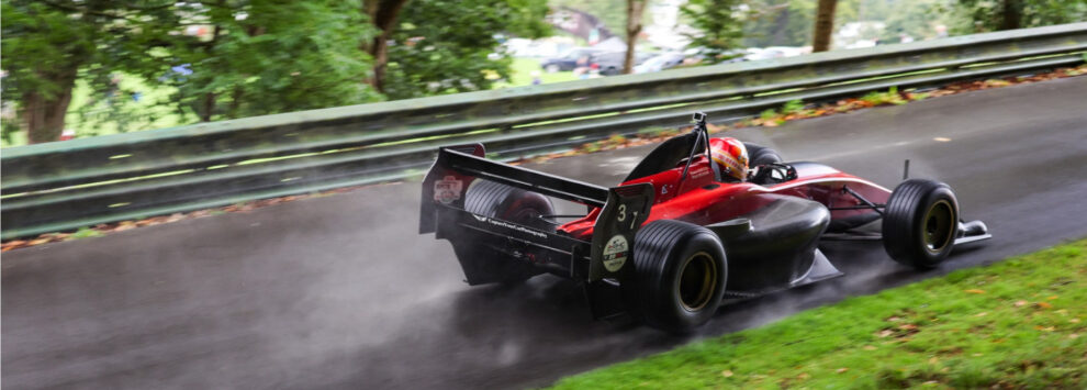 A red single seater racing car climbing a wet, tree lined hillclimb course past a steel barrier