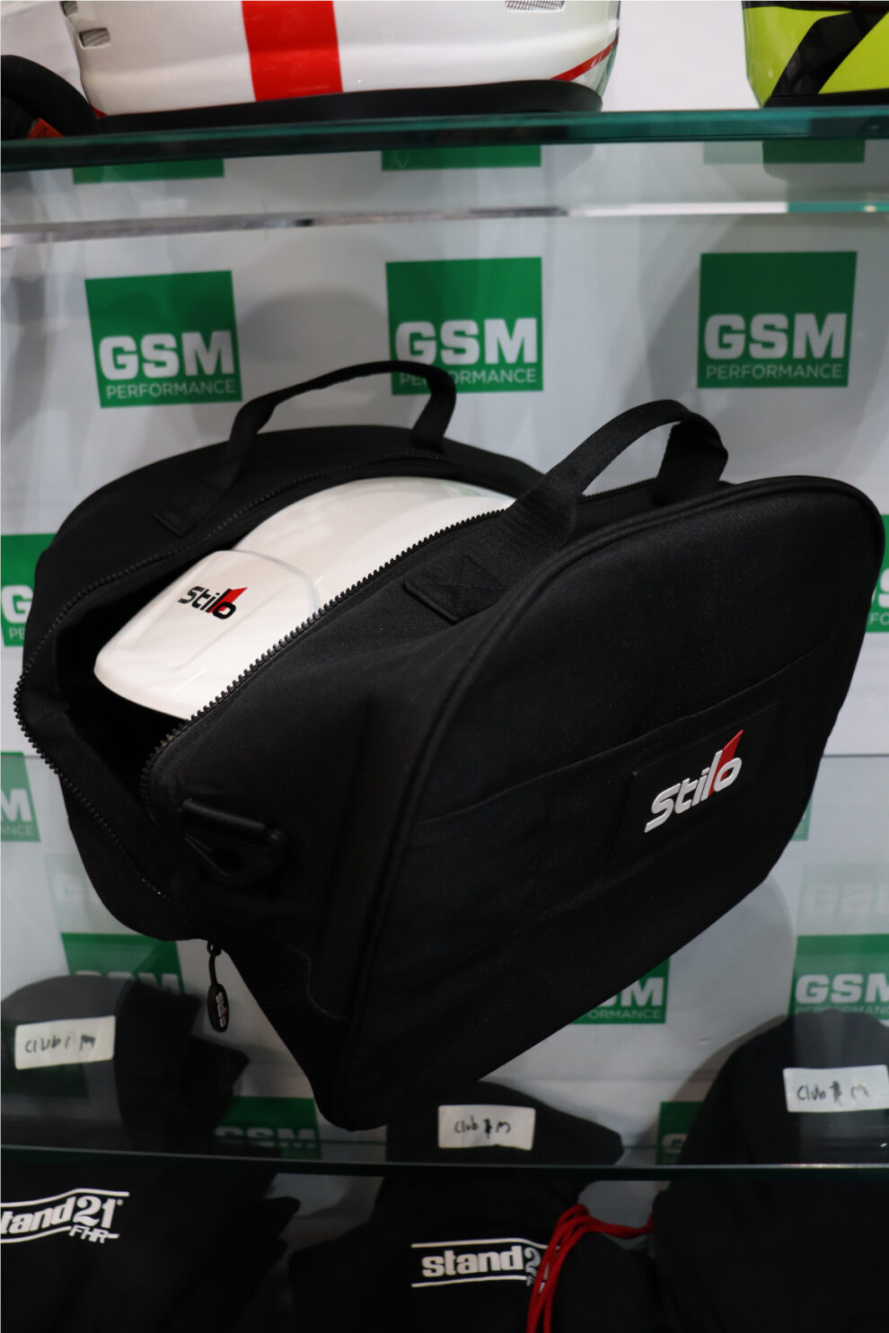 A white Stilo helmet packed inside a black Stilo helmet and FHR bag on a showroom shelf, with black balaclavas on the shelf below