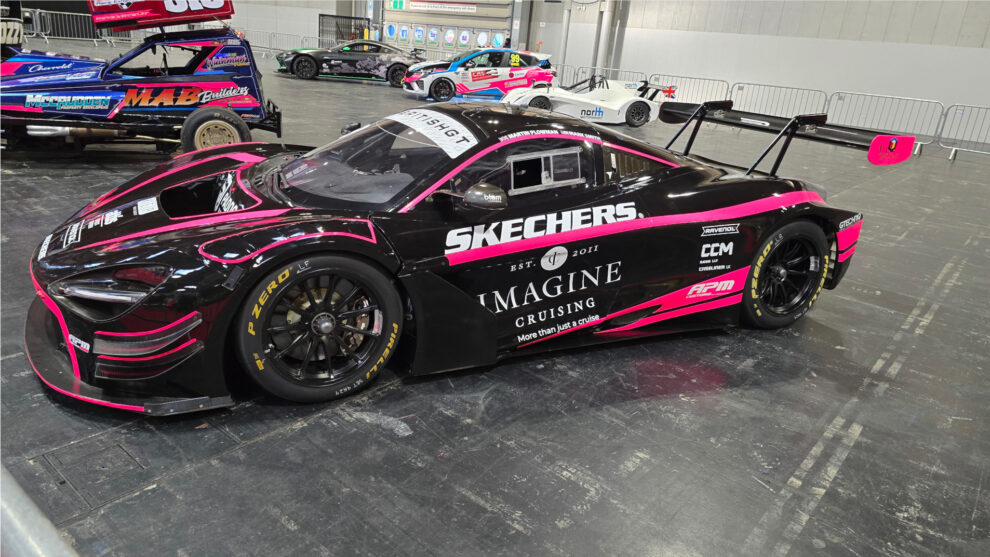 A black and pink McLaren GT3 race car with a large rear wing on display at Autosport International 2025, with a stock car behind