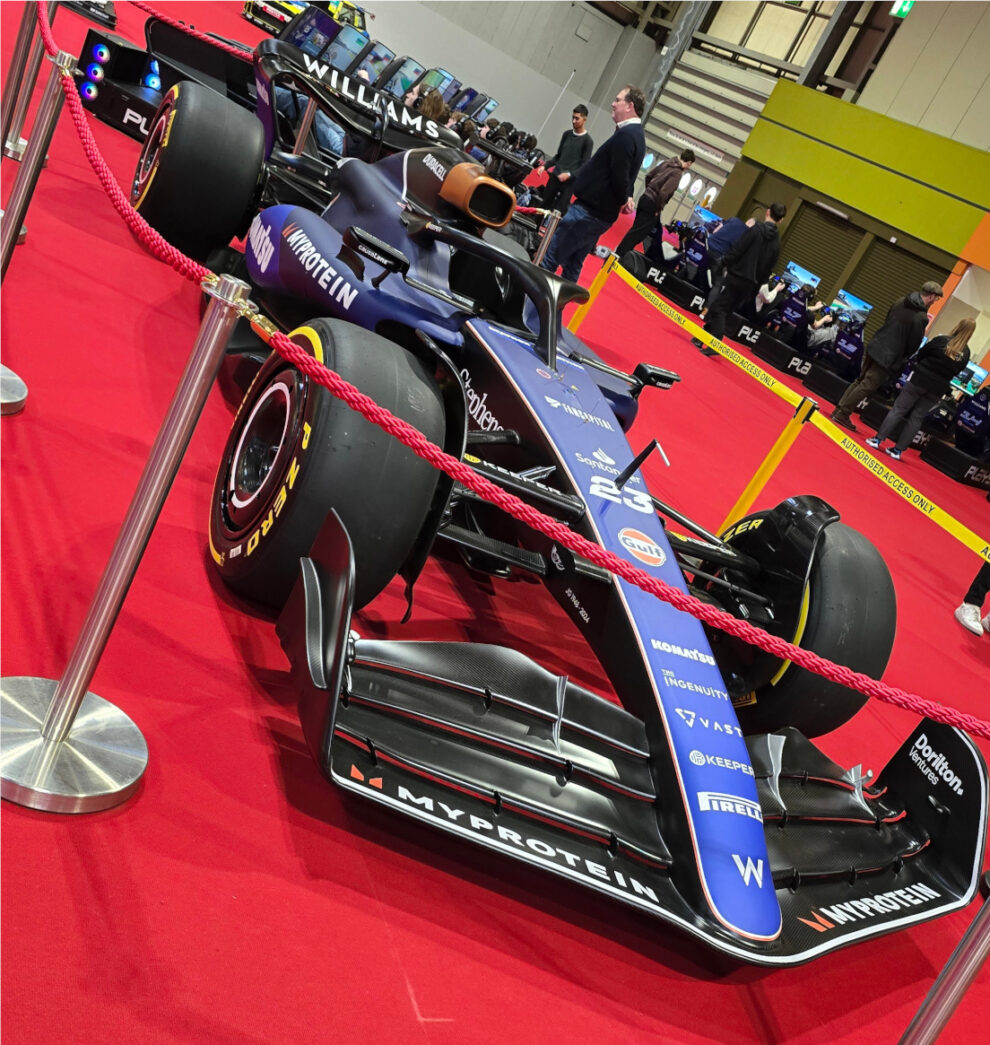 A Williams Formula 1 car in dark blue livery with the number 23 behind a rope barrier on red carpet at Autosport International 2025