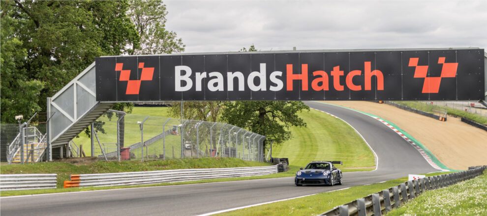 A blue Porsche 911 GT3 RS driving under the Brands Hatch bridge with the track climbing behind it