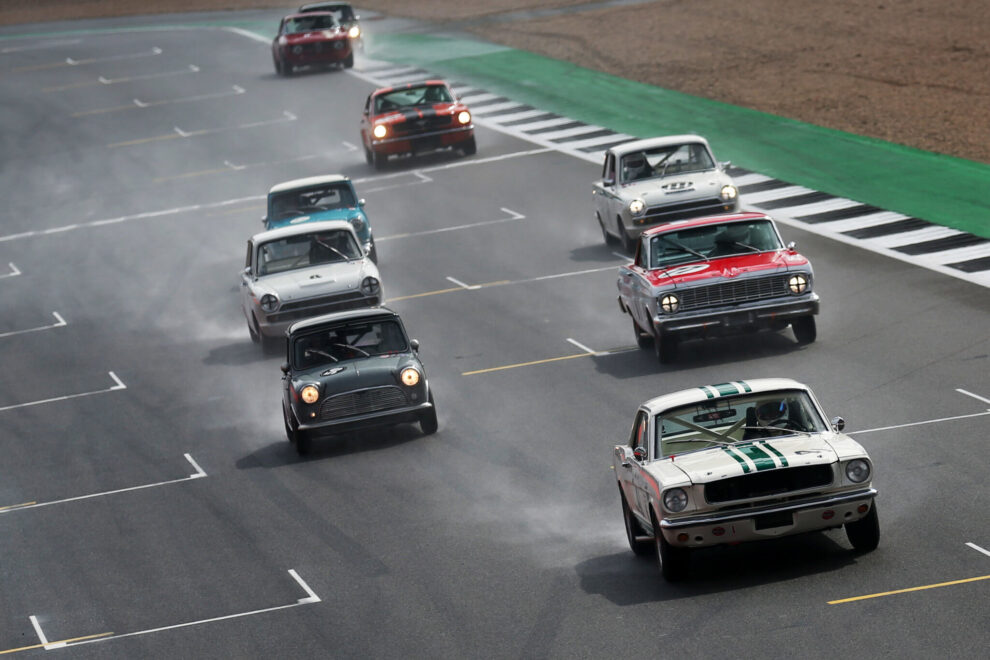 A pack of historic saloon racing cars led by a white Ford Mustang, with a Mini and other historic saloons behind, racing on a wet circuit