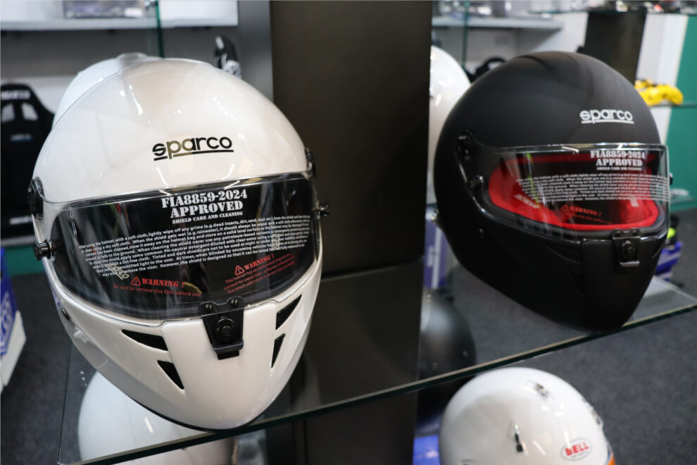A white and a matt black Sparco Stealth RF helmet with FIA 8859-2024 visor stickers on a glass shelf in the GSM Performance showroom
