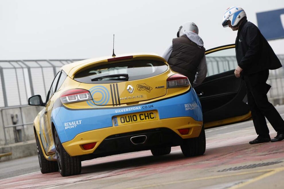A yellow and blue Renault Megane RS tuition car in the Silverstone pit lane with a helmeted driver and an instructor at the open door