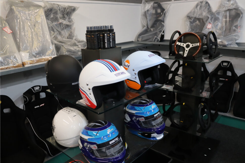 The Martini Racing and Gulf J-Pro helmets on a glass display stand in the GSM Performance showroom, with a black J-Pro, Zamp helmets and steering wheels nearby