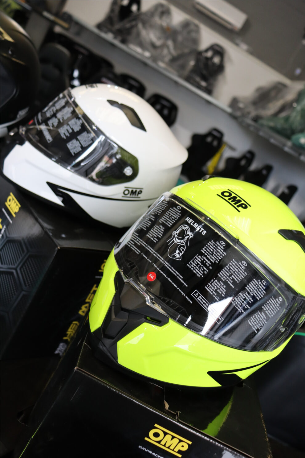 A fluorescent yellow OMP full face helmet and a white OMP helmet on their boxes in the GSM Performance showroom, with seats on the shelves behind