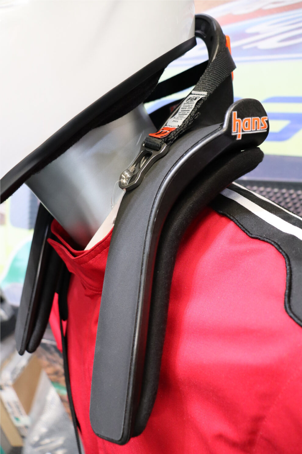 Close view of a HANS IV device on the mannequin, showing the tether, its FIA label and the clip that attaches to the helmet post