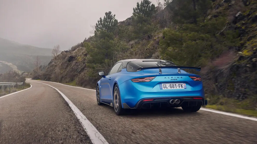 The rear of a blue Alpine A110 R with a rear wing and centre exhaust, driving along a winding hillside road