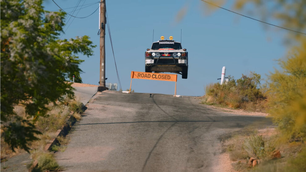 The Subaru Brataroo 9500 Turbo in the air over the crest of a road beside an orange Road Closed sign