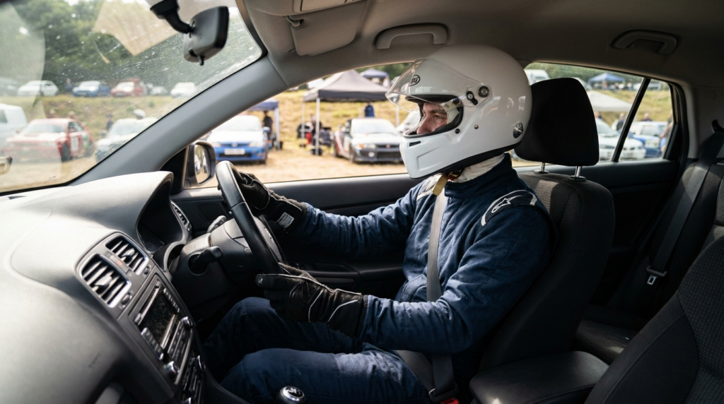 Illustration of a driver wearing a race suit, balaclava, gloves and full-face helmet in a standard road car seat with a factory three-point seatbelt