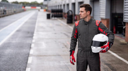Racing driver in an Alpinestars GP Race V3 suit holding a white helmet with a dark visor in a UK circuit pit lane