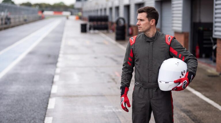 Racing driver in the Alpinestars GP Race V3 race suit in Anthracite and Red standing in a UK circuit pit lane, holding a plain white helmet