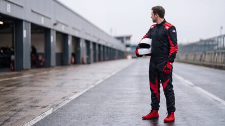 Racing driver in the OMP One Evo X race suit in Black & Red looking down a circuit pit lane, plain white helmet under one arm